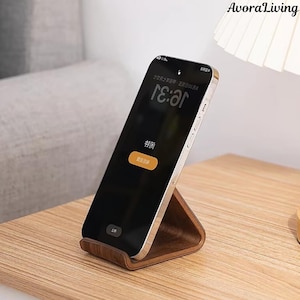 Könnte beinhalten: Dunkelbrauner Handyhalter aus Holz mit einem Smartphone, das einen Sperrbildschirm anzeigt. Der Ständer hat ein geschwungenes Design. Auf dem Bildschirm ist die Uhrzeit 7:31 zu sehen. Das Telefon ist silber und schwarz.