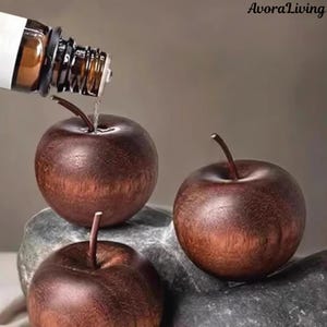 May include: Three wooden apple-shaped ornaments with stems are displayed on a grey stone surface. A brown bottle is pouring liquid onto one of the apples. The apples have a dark brown, polished finish.