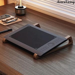 Könnte beinhalten: Ein graues digitales Zeichentablett mit schwarzem Bildschirm, das auf einem Holzständer ruht. Das Tablet hat Steuertasten und ein kreisförmiges Zifferblatt. Ein Stift befindet sich in der Nähe. Der Markenname "AvoraLiving" ist sichtbar.