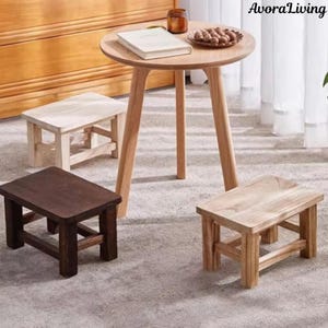 May include: A set of wooden furniture, featuring a small round table and three small stools. The table has a light wood finish, with a book and snacks on top. The stools are in various shades of brown and natural wood.