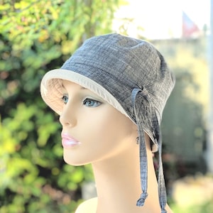 May include: A gray cloche hat with a white brim and matching ties. The hat is made of a textured fabric and has a rounded crown. The ties are knotted at the ends. The hat is displayed on a mannequin.