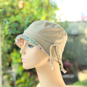 May include: A khaki-colored sun hat with a patterned brim featuring blue, green, and pink floral designs. The hat has a small brim and ties on the sides. A small charm hangs from the side.