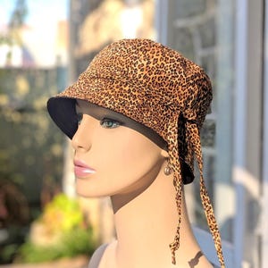 May include: A leopard print bucket hat with a black brim and matching ties. The hat features a brown and tan animal print pattern. The ties hang down the side of the hat. The hat is displayed on a mannequin.
