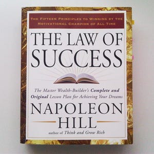 May include: A hardcover book titled "The Law of Success" by Napoleon Hill. The cover features the title in large black letters, an open book illustration, and the author's name. The book's cover has a gold and brown decorative border.