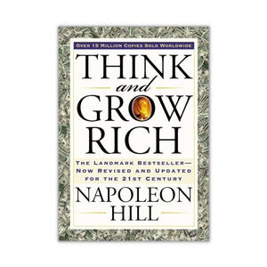 Könnte beinhalten: Buchcover von "Think and Grow Rich" von Napoleon Hill. Der Titel ist in großen schwarzen Buchstaben mit einer goldfarbenen Grafik gehalten. Der Einband ist von einem Geldrahmen umgeben.