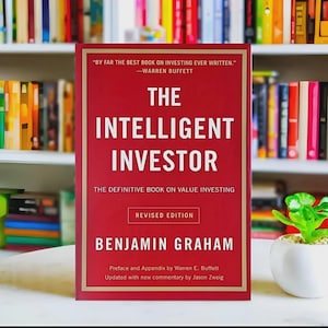 Op de afbeelding: Een rood boek met de titel "The Intelligent Investor" met witte letters. Het boek is van Benjamin Graham en bevat een citaat van Warren Buffett. Het boek ligt op een tafel met een boekenplank op de achtergrond.