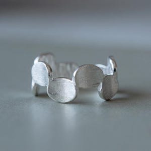 May include: A silver ring with a unique design. The ring features a series of connected, rounded shapes, creating a textured appearance. The metal has a matte finish, reflecting light softly. This piece of jewelry is suitable for everyday wear.