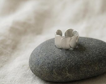 May include: A silver ring with a distinctive design, composed of several rounded, overlapping elements. The ring is presented on a grey stone, with a neutral fabric backdrop. The ring has a matte finish, reflecting soft light.