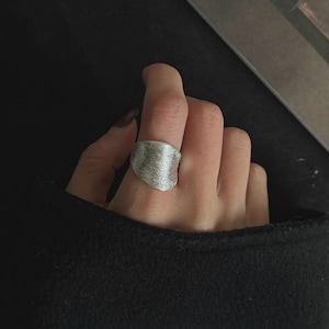 May include: A silver-colored ring with a textured surface. The ring has an irregular, organic shape and is worn on a finger. The background is dark, emphasizing the ring's metallic appearance. The ring is a piece of jewelry.