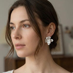 May include: Silver-colored flower-shaped stud earrings. The earrings have a textured, organic design, resembling a blooming flower. The earrings are worn on the ear of a person with brown hair and fair skin. The background is blurred.