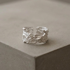 Peut inclure: Une bague argentée au design texturé et organique. La bague a un design ouvert, permettant de légers ajustements de taille. La surface de la bague présente une texture rugueuse et inégale, lui conférant un aspect unique et artisanal. La bague est présentée sur une surface grise neutre.