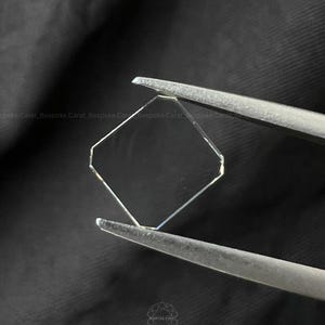 May include: A clear, colourless, hexagonal-cut gemstone held by metal tweezers. The stone features flat, polished facets and a geometric shape. The text 'Bespoke Carat' is visible in the background and as a small logo at the bottom centre.