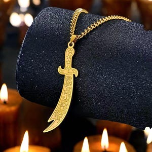 Puede incluir: Collar con colgante dorado con un diseño de daga curvada. El colgante tiene detalles intrincados y escritura árabe. La cadena es de color dorado a juego. El collar se muestra sobre un fondo oscuro y texturizado.