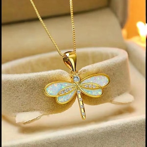 Peut inclure: Collier pendentif libellule doré. Le pendentif libellule présente des ailes opalescentes avec des accents de cristaux clairs et un diamant central. Le collier est présenté dans un écrin beige, mettant en valeur le design et le savoir-faire.