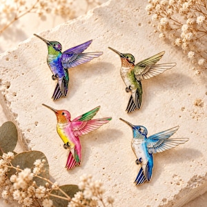 May include: Four hummingbird-shaped enamel pins featuring vibrant colour gradients. One pin is purple and green, one is orange and green, one is pink and yellow, and one is blue and white. Each pin displays detailed wing feathers and a long beak.