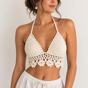 May include: A cream-colored crochet halter top featuring a deep V-neckline, thin neck straps, and a decorative hemline composed of repeating flower motifs. The bodice is constructed with a dense stitch pattern, transitioning into an openwork design.