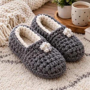 May include: A pair of dark grey crochet slippers featuring a thick, cream-coloured interior lining and a small, cream-coloured crocheted flower embellishment on the top of each slipper. The slippers have a chunky, textured yarn construction.