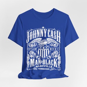 May include: A royal blue t-shirt featuring a white graphic design. The design includes the text "JOHNNY CASH" above an eagle emblem, with "OUTLAW MUSIC" on either side. Below, it reads "MAN IN BLACK NASHVILLE TENNESSEE."