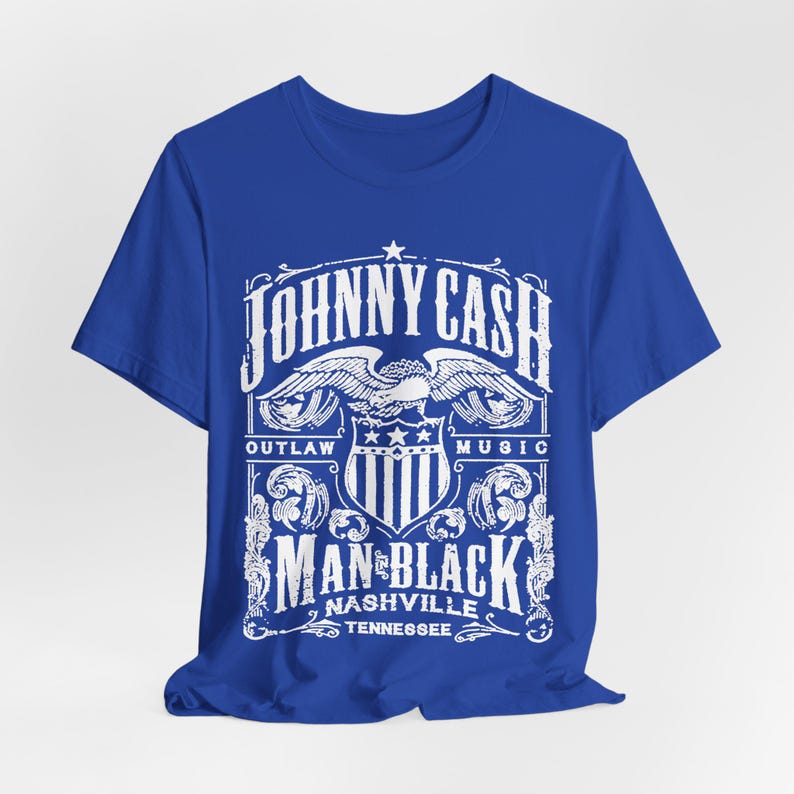 May include: A royal blue t-shirt featuring a white graphic design. The design includes the text "JOHNNY CASH" above an eagle emblem, with "OUTLAW MUSIC" on either side. Below, it reads "MAN IN BLACK NASHVILLE TENNESSEE."