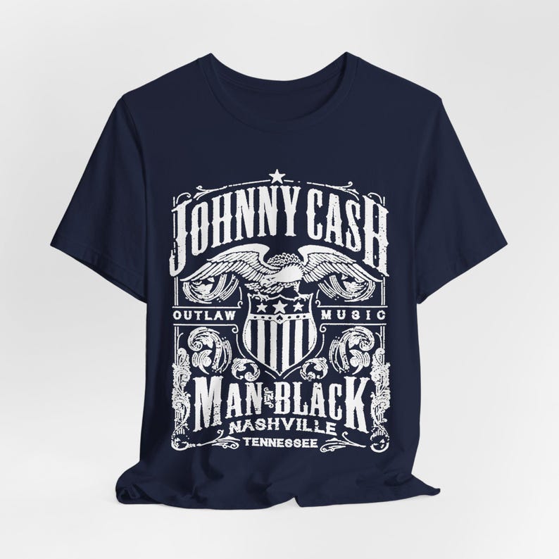 May include: Navy blue t-shirt featuring a white graphic design. The design includes the text "JOHNNY CASH" above an eagle emblem, with "OUTLAW MUSIC" on either side. Below, it reads "MAN IN BLACK NASHVILLE TENNESSEE".