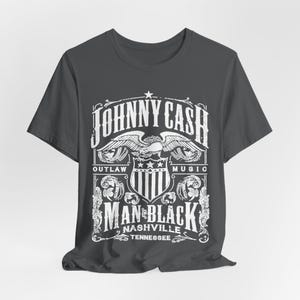 May include: Dark gray t-shirt featuring a white graphic design. The design includes the text "JOHNNY CASH" above an eagle emblem, with "OUTLAW MUSIC" on either side. Below, it reads "MAN IN BLACK NASHVILLE TENNESSEE".