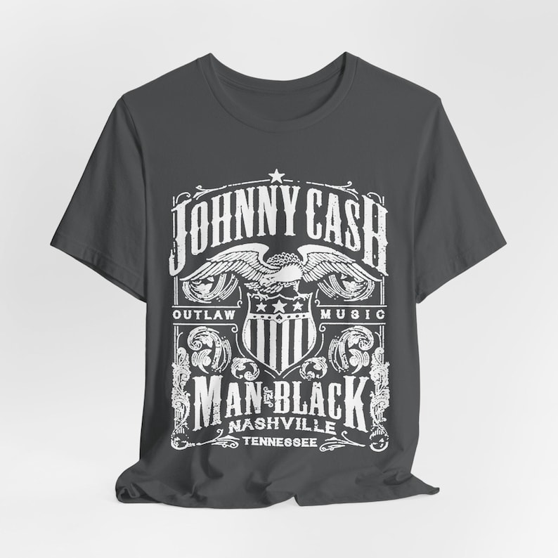 May include: Dark gray t-shirt featuring a white graphic design. The design includes the text "JOHNNY CASH" above an eagle emblem, with "OUTLAW MUSIC" on either side. Below, it reads "MAN IN BLACK NASHVILLE TENNESSEE".