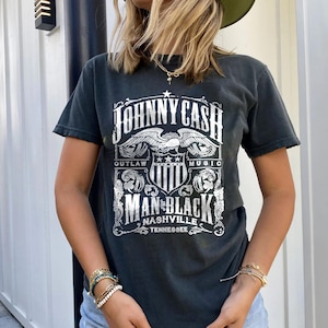 May include: Black t-shirt featuring a graphic design with the text "JOHNNY CASH MAN IN BLACK NASHVILLE TENNESSEE". The design includes an eagle and shield motif. The shirt has a vintage, worn look.