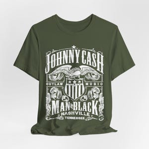 May include: Olive green t-shirt featuring a white graphic design. The design includes the text "JOHNNY CASH" above an eagle emblem, with "OUTLAW MUSIC" and "MAN IN BLACK NASHVILLE TENNESSEE" below. The shirt is made of a soft material.