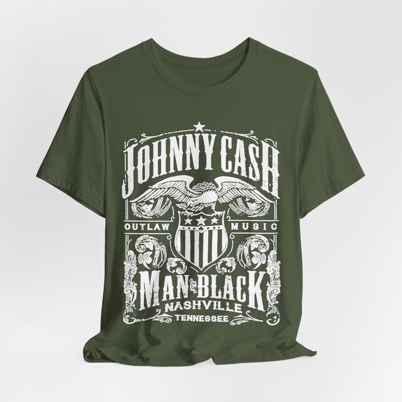 May include: Olive green t-shirt featuring a white graphic design. The design includes the text "JOHNNY CASH" above an eagle emblem, with "OUTLAW MUSIC" and "MAN IN BLACK NASHVILLE TENNESSEE" below. The shirt is made of a soft material.