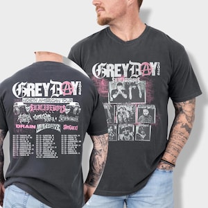 May include: A charcoal gray V-neck t-shirt featuring a graphic print with the text GREY DAY 2026, a pink anarchy symbol, and a collage of black and white photographs. The back displays a tour schedule with city names and dates in white text.