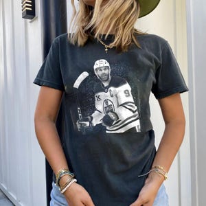 May include: Dark gray t-shirt featuring a black and white image of a hockey player holding a hockey stick. The player is wearing a white and blue jersey with the number 9. The shirt has a distressed, vintage look.