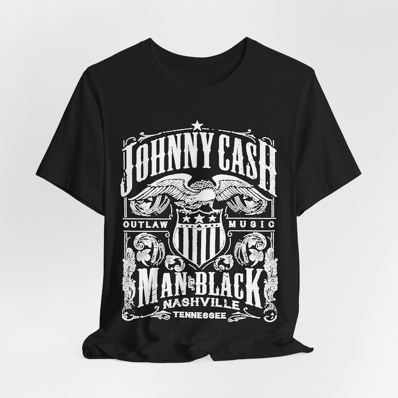 May include: Black t-shirt featuring a white graphic design. The design includes the text "JOHNNY CASH" above an eagle emblem, with "OUTLAW MUSIC" on either side. Below, it reads "MAN IN BLACK NASHVILLE TENNESSEE."