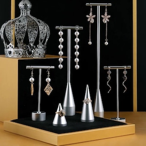 May include: An assortment of jewelry displayed on silver stands and ring cones. The earrings include pearl drops, floral designs, and geometric shapes. A decorative crown sits in the background, and the display is set on a black velvet surface.