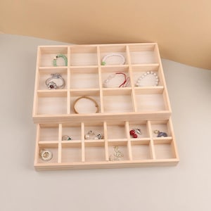 May include: Two-tiered, light-colored wooden jewelry organizer with multiple compartments. The top tier holds rings and bracelets, including a green beaded bracelet and a silver ring with a floral design. The bottom tier contains earrings and other small jewelry items.