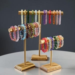 May include: A display of beaded bracelets in various colors, including pink, blue, green, and orange, are showcased on gold-colored stands. The bracelets are made of small, round beads. The stands have a wooden base and a horizontal bar to hold the bracelets.