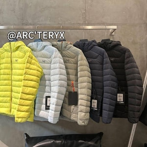May include: A row of hooded puffer jackets in various colors, including yellow, light blue, gray, navy, and black, hanging on a metal rack. The jackets have a quilted design and a small logo on the chest. The text "@ARC'TERYX" is visible.