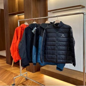 May include: A clothes rail holds a selection of jackets. There's a bright orange hooded jacket, a black jacket, a blue and black jacket, and a black quilted jacket with a logo. The jackets are suspended on wooden hangers.
