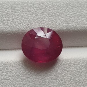 May include: A faceted, oval-shaped ruby gemstone. The gem is a deep, rich red colour and appears to be set against a white, textured surface. The cut of the stone creates multiple facets that reflect light.