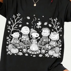 以下が含まれることがあります： キャンプファイヤーを囲むピーナッツのキャラクターたちが描かれた、黒の半袖Tシャツ。白い線画でスヌーピー、チャーリー・ブラウン、仲間たちが丸太に座り、カボチャやランタン、秋の落ち葉がデザインされています。
