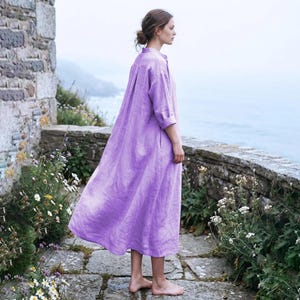 Puede incluir: Un vestido de manga larga y largo hasta el tobillo en un vibrante color lavanda. La prenda presenta un cuello abotonado, una silueta holgada y fluida, y mangas con puños. La tela tiene un tejido texturizado visible y el diseño incluye un dobladillo recto simple que cae por debajo de la pantorrilla, proporcionando un ajuste relajado y cómodo.