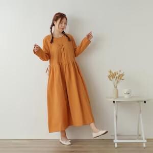 May include: An orange long-sleeved dress with a gathered waist and a flowing skirt. The dress has a round neckline and is worn with white flats. A small white table with a vase of dried flowers is in the background.