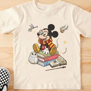 May include: Cream-colored t-shirt featuring a cartoon Mickey Mouse character wearing glasses and a scarf, sitting on books. The design includes a small owl, a wand, and a golden snitch. The shirt is on a wooden surface.