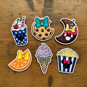 May include: A collection of colorful embroidered patches featuring food and drink designs. Includes a blue polka-dot drink with a cherry, a chocolate chip cookie, a crescent moon, an orange slice, an ice cream cone, and a popcorn container.