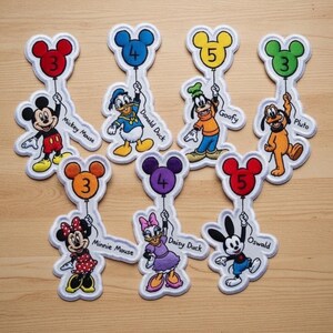 May include: Embroidered patches featuring Disney characters holding balloons with numbers. Characters include Mickey Mouse, Donald Duck, Goofy, Pluto, Minnie Mouse, Daisy Duck, and Oswald. Each patch has a white border and the character's name.