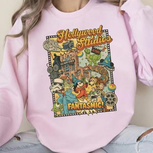 Può includere: Felpa rosa chiaro con una grafica colorata delle attrazioni e dei personaggi di Hollywood Studios. Il design include il testo "Hollywood Studios" e "Fantasmic!" con immagini di una fotocamera, una bobina di film e personaggi dei cartoni animati.