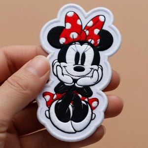 May include: Embroidered patch featuring Minnie Mouse. The patch shows Minnie Mouse in a seated pose, with a red and white polka dot bow, black ears, and a smiling face. The patch has a white border.