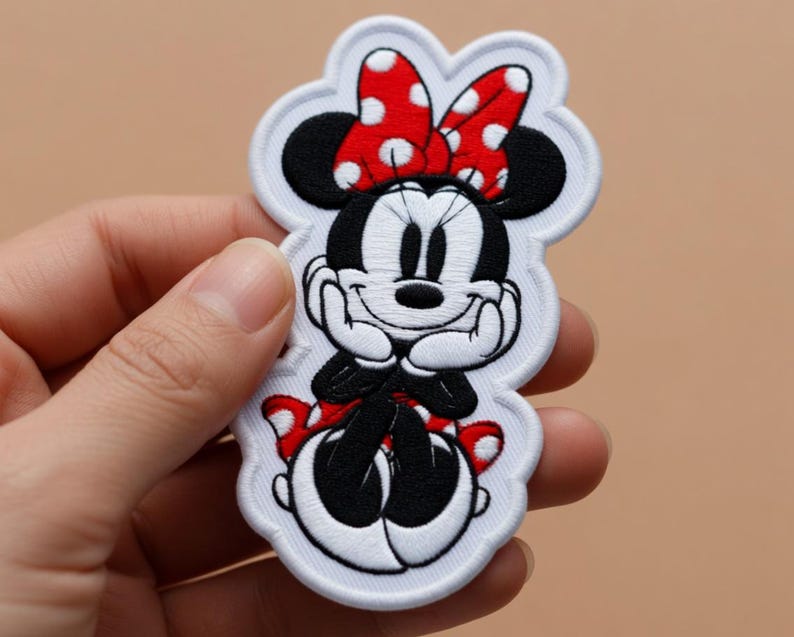 May include: Embroidered patch featuring Minnie Mouse. The patch shows Minnie Mouse in a seated pose, with a red and white polka dot bow, black ears, and a smiling face. The patch has a white border.