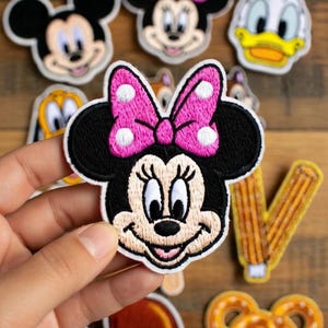 May include: Embroidered patches featuring cartoon characters, including Mickey Mouse, Minnie Mouse with a pink bow, and Donald Duck. Other patches include a churro, a pretzel, and a turkey leg. The patches are on a wooden surface.