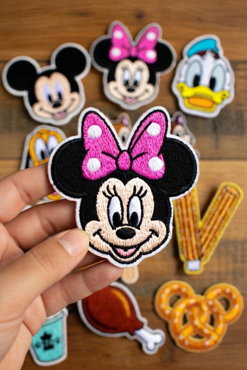 May include: Embroidered patches featuring cartoon characters, including Mickey Mouse, Minnie Mouse with a pink bow, and Donald Duck. Other patches include a churro, a pretzel, and a turkey leg. The patches are on a wooden surface.