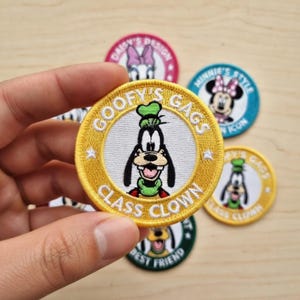 Puede incluir: Parches bordados con personajes de dibujos animados. El parche principal muestra a Goofy con el texto "GOOFY'S GAGS CLASS CLOWN". Otros parches incluyen a Daisy Duck, Minnie Mouse y el texto "BEST FRIEND".